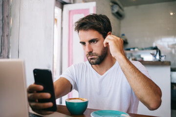 Thoughtful young bearded man using smartphone while drinking coffee in cafe