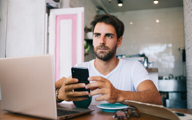 Handsome caucasian male blogger holding mobile phone thinking about idea for publication working remotely, pensive young hipster guy freelancer using modern device and 4G internet connection