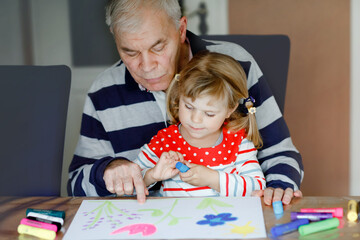 Cute little baby toddler girl and handsome senior grandfather painting with colorful felt pens and pencils at home. Grandchild and man having fun together, creative family.