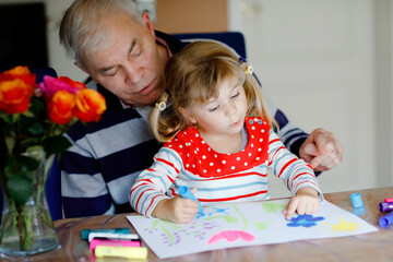 Cute little baby toddler girl and handsome senior grandfather painting with colorful felt pens and pencils at home. Grandchild and man having fun together, creative family.