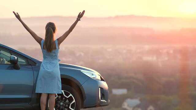 Happy young woman driver with outstretched up hands enjoying warm summer evening standing beside her car. Travelling and vacation concept.