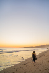 Young woman walking along the golden sandy beach and fog with shoes in hand on Falesia Beach at sunset on the Atlantic Ocean. Olhos de Agua, Algarve, Portugal.