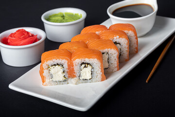 Sushi Sets Uramaki, California, Philadelphia, on a white plate. Nearby ginger and wasabi. Soy sauce in a white bowl. On a dark background.