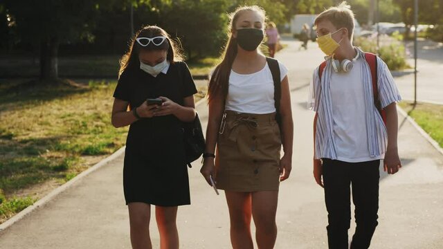 Schoolchildren, boy and two girls, wearing protective face masks and backpacks are walking down the street, taking selfies, talking and laughing