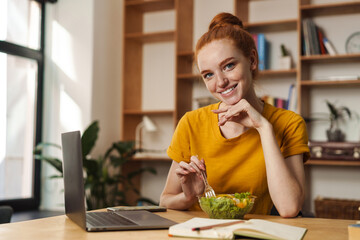 Image of smiling redhead girl eating salad while working with laptop