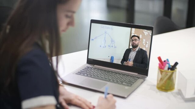 Girl participates in the training program. Woman is watching an online lesson, making notes while looking at the computer screen following her teacher Learning at home remotely.