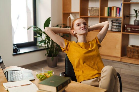 Image of relaxed redhead girl using earphones while working with laptop