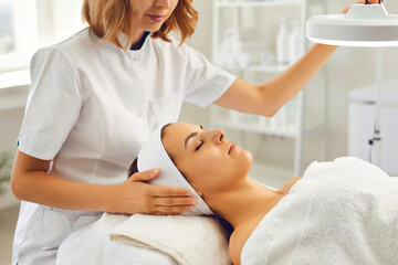 Cosmetologist or dermatologist directing lamp to womans face for facial treatment