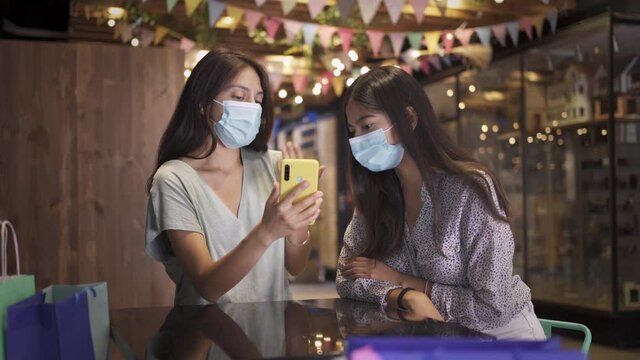 Two young women spending time together while chatting and looking at the phone  during the coronavirus pandemic. Girls having fun in a bar in the evening wearing sanitary masks to prevent the virus. 