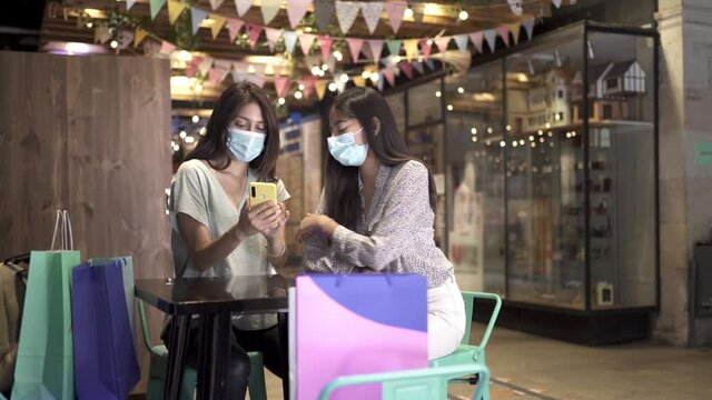 Two young women friends are chatting and looking at the phone after shopping during the coronavirus pandemic. Two girls with sanitary protection masks in a cafe of a mall during the covid-19 crisis. 