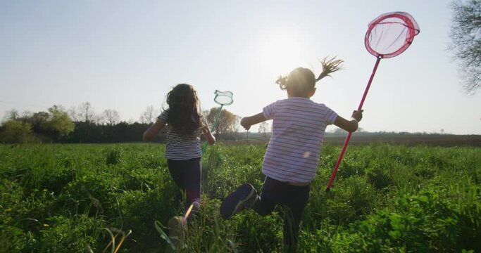 Authentic shot of two carefree happy little girls are having fun to run with butterfly net on a countryside green fields in a sunny day. Concept: happiness, freedom, childhood, nature, authenticity