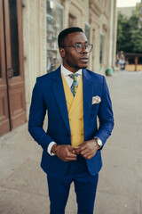 Handsome Afro-American fashion model in a blue suit and yellow waistcoat posing near house.