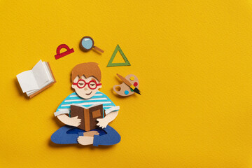 Smiling guy in glasses reading educational schoolbook. Book, protractor, magnifier, triangle ruler palette of watercolors above pupil. Education concept. Hand cut paper illustration with copy space