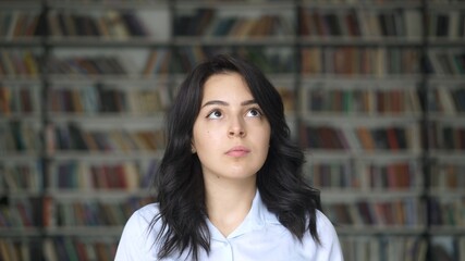 Young woman thinking, background of blurred bookshelves in a big library. Close up of asian student in white shirt thinking over, choosing a book