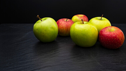 Three ripe fresh green apples and tree red apples on black table and dark background. Free copy space. School snacks or organic food concept