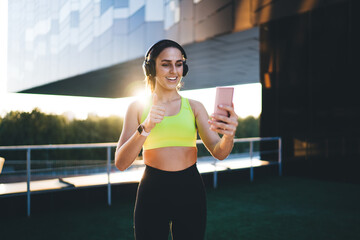 Carefree Caucasian female jogger in active wear posing for smartphone camera making selfie during sunny morning, smiling sports woman in headphones fitness influencer using cellular for web seminar