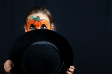 a girl in pumpkin makeup for Halloween hides behind a black hat in her hands on a black background, isolated.