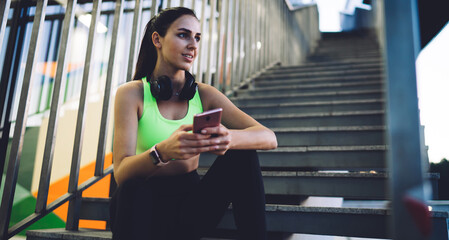 Fototapeta premium Pensive athletic woman with smartphone sitting on steps and thinking over read message