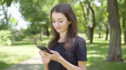A young Turkish woman listens to music with earphones on a smartphone and smiles in a park on a sunny day