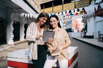 Cheerful female students enjoying friendly meeting for social networking via touch pad, happy hipster girls with education textbook using digital tablet with 4g wireless for watching web video