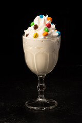 Classic milkshake decorated with sweets and whipped cream in a transparent glass on a black background