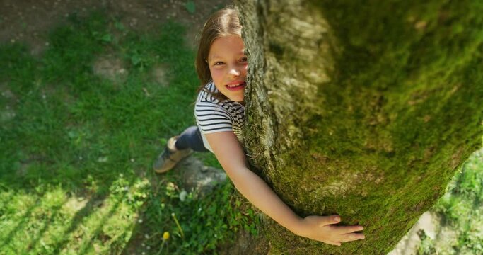 Authentic shot of a happy little girl is hugging a tree trunk and smiling in camera on a sunny day. Concept: love for nature, connection with nature, happiness, childhood, ecology, deforestation