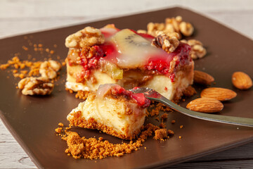 A slice of fruit cheesecake served on a brown porcelain plate with walnuts and almonds. The cake has raspberry and kiwi fruits which is finished with custard icing. A metal fork separates a bite size.