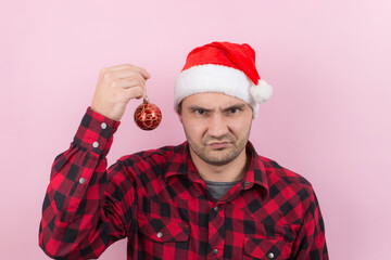 Unhappy, upset man in red hat, holds Christmas decoration with disgust on his face. Negative human emotions christmas. Pink background, copy space.