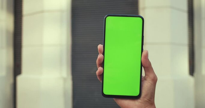 Close up view of female hand holding smartphone and pressing on green blank screen. Concept of mockup and chroma key. Closed building at background.