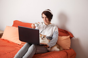 A person holding a cat and laptop. a home schooling and work at home. A woman in white headphones, in home clothes with a cat on the bed sits and looks at the laptop.