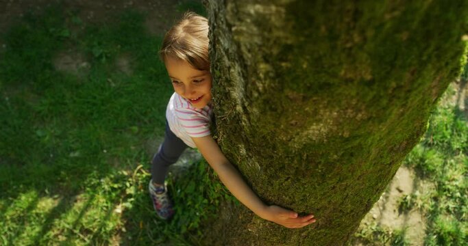 Authentic shot of a happy little girl is hugging a tree trunk and smiling in camera on a sunny day. Concept: love for nature, connection with nature, happiness, childhood, ecology, deforestation