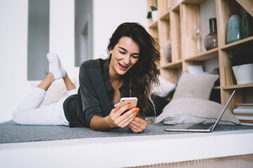 Cheerful caucasian female laughing at funny message getting on mobile phone working remotely on laptop computer, smiling woman having fun at free time in home interior using modern devices and wifi