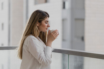 Happy young woman sniffs cup of tea or coffee with blurred city background. Girl dressed in white shirt drinking tea at balcony. Relaxing breathing fresh in the morning. Beautiful life in the city