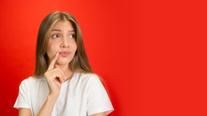 Choosing, thoughts. Portrait of young caucasian woman isolated on red studio background with copyspace. Blonde female model. Concept of human emotions, facial expression, sales, advertising, youth