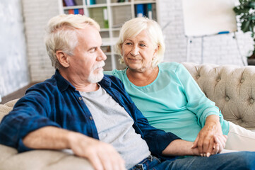 Senior wife looks at her senior husband with support and empathy. Elderly gray-haired couple sits on the sofa at home