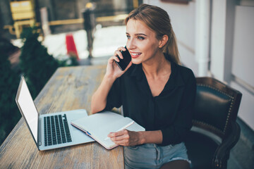 Happy female student receiving positive cellphone call sitting outdoors with laptop device and education textbook for e learning, prosperous freelancer communicate via smartphone and smiling
