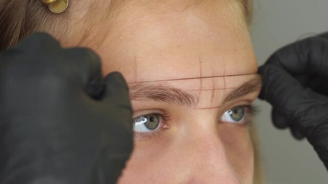 Professional styling, correction and shaping procedures of female eyebrows in beauty salon. Closeup view of client face and stylist's hands working with special thread to mark new contour of brows.