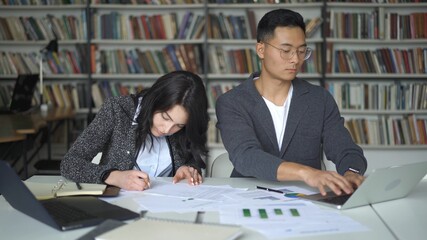 High school students working in library after classes, with paper, charts and laptops. Young man and woman sitting doing notes on background of bookshelves