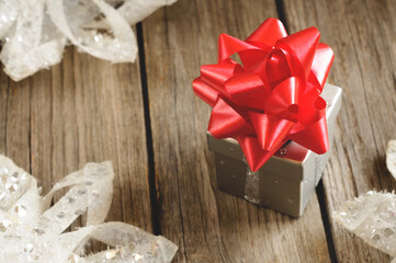 Small gift box with a large red bow on a wooden background with Christmas decorations. Blurred background and foreground. Selective focus on bow