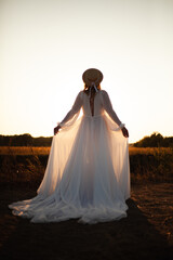 Happy woman standing with her back on sunset in nature .
Woman in a long white dress with a hat on her head