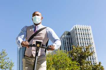 Black businessman in formal clothes using an electric scooter to move around the city. He is wearing a medical mask. New normal and social distancing concept.