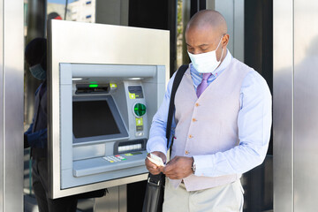 African American entrepreneur withdrawing cash at an ATM. He is wearing a medical mask as a protective measure during the Covid-19 pandemic.