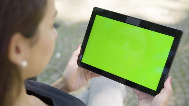 A Turkish woman looks at a tablet with green screen - horizontal position - focused closeup from behind - she sits on a bench outside