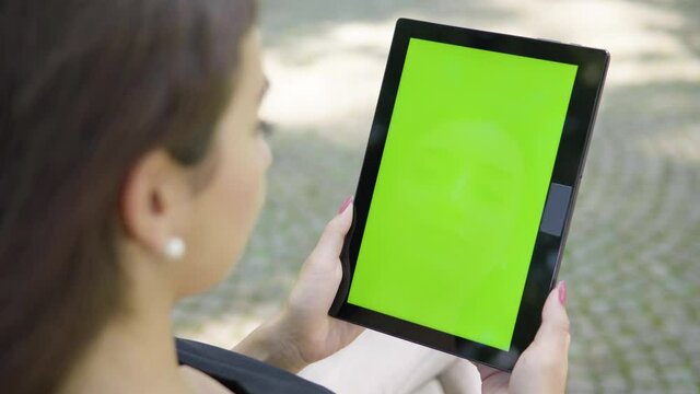 A Turkish woman looks at a tablet with green screen - vertical position - focused closeup from behind - she sits on a bench outside