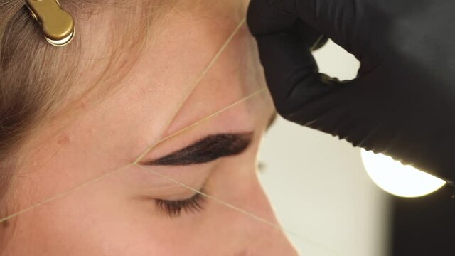 Professional styling, correction and lamination procedures of female eyebrows in beauty salon. Closeup view of client face and stylist's hands working in gloves. Master removes hair using thread.