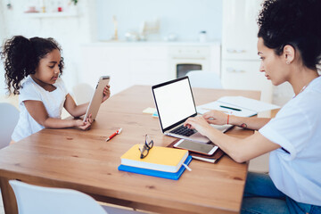 Young modern mother typing on laptop with girl relaxing near