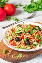 Pearl barley salad with vegetables in a plate close-up. Boiled pearl barley with tomatoes, cucumbers, olives, parsley and basil. Vegan and vegetarian food.