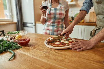 Cropped shot of man, cook cutting hot freshly baked pizza on the table while his girlfriend drinking wine. Couple making pizza together. Hobby, lifestyle