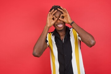 African american man with braids wearing casual shirt over isolated red background doing ok gesture like binoculars sticking tongue out, eyes looking through fingers. Crazy expression.