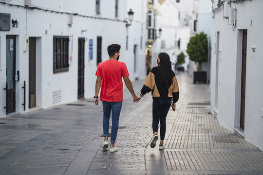 Shallow Focus Of A Young Couple With Facemasks Holding Hands While Walking Down An Alley